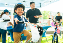 NeighborHealth promotes safe summer riding through its Annual Childrenโs Bike Safety Day Smiling young boy wearing a helmet rides a white bike at an outdoor event, others in background under a canopy