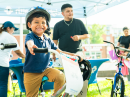 NeighborHealth promotes safe summer riding through its Annual Childrenโs Bike Safety Day Smiling young boy wearing a helmet rides a white bike at an outdoor event, others in background under a canopy