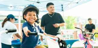 NeighborHealth promotes safe summer riding through its Annual Childrenโs Bike Safety Day Smiling young boy wearing a helmet rides a white bike at an outdoor event, others in background under a canopy
