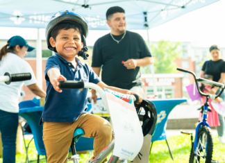 NeighborHealth promotes safe summer riding through its Annual Childrenโs Bike Safety Day Smiling young boy wearing a helmet rides a white bike at an outdoor event, others in background under a canopy