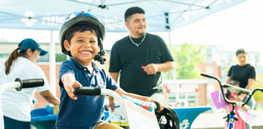 NeighborHealth promotes safe summer riding through its Annual Childrenโs Bike Safety Day Smiling young boy wearing a helmet rides a white bike at an outdoor event, others in background under a canopy