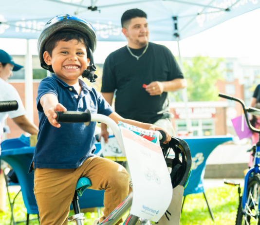 NeighborHealth promotes safe summer riding through its Annual Childrenโs Bike Safety Day Smiling young boy wearing a helmet rides a white bike at an outdoor event, others in background under a canopy