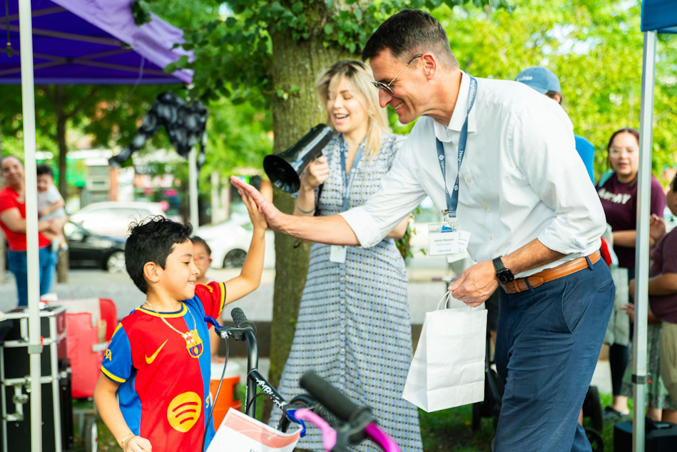 Man in a white shirt high-fives a smiling boy in a red and blue soccer jersey at an outdoor community event near bikes and tents.